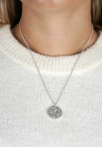 Silverfärgad halskedja med en texturerad, oregelbunden hängande del på en tunn kedja, bärs över en vit stickad tröja.