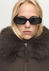Chaqueta acolchada marrón con un oscuro cuello de piel sintética, cierre de cremallera, y gafas de sol negras rectangulares y delgadas. Textura suave en general.