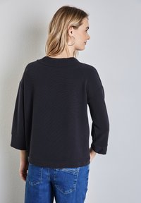 Svart, texturerad långärmad topp med avslappnad passform och rund halsringning, tillsammans med blå jeans, sedd bakifrån.