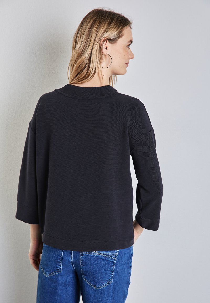 Svart, texturerad långärmad topp med avslappnad passform och rund halsringning, tillsammans med blå jeans, sedd bakifrån.