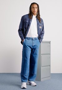 Blue and white striped button-up shirt, white T-shirt, loose blue denim jeans, and white sneakers, standing next to a gray filing cabinet.