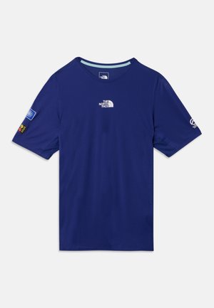 Mand stående iført en blå kortærmet skjorte, orange shorts, orange kasket, orange sokker og hvide løbesko mod en ensfarvet baggrund.