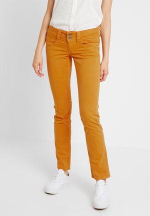 Mujer con pantalones ajustados color mostaza, zapatillas blancas y camisa sin mangas blanca abotonada, de pie sobre un suelo gris.