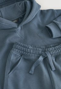 Jasnoniebieska bluza z kapturem i dopasowane dresy wykonane z miękkiej mieszanki bawełny. Posiadają ściągacze przy mankietach oraz elastyczną talię ze sznurkiem do regulacji.