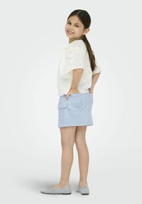 Witte eyelet blouse, lichtblauwe gestreepte cargorok, grijze ballerina's. Kind staat zijwaarts, handen in de zakken, met lang donker haar.