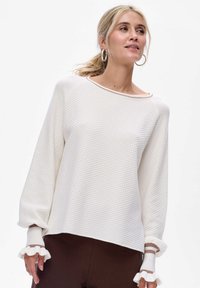 Witte gebreide trui met een brede halslijn, lange mouwen met rimpeldetails en contrasterende bruine accenten op de manchet.