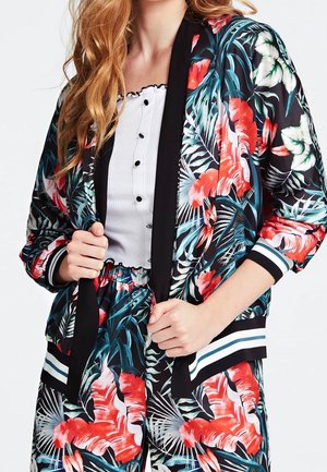 Kvinde iført en jakke med blomsterprint og matchende bukser med røde, hvide og grønne blade over en hvid knapbluse.