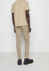 Beżowa koszulka z krótkim rękawem i dopasowane beżowe spodnie o kroju slim. Czarne botki z teksturowanym wykończeniem. Neutralne tło.