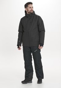 Svart vattentät jacka med dragkedja och huva, i kombination med svarta isolerade byxor som har elastiska muddar och förstärkta detaljer.