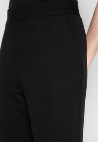 Gros plan sur un pantalon noir sur mesure porté à la taille, montrant les poches latérales et une partie du bras d'une personne sur un fond clair.