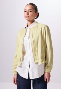 Giacca bomber gialla chiara con polsini e orlo elasticizzati, realizzata in tessuto liscio. Indossata sopra una camicia bianca a maniche lunghe e jeans blu.