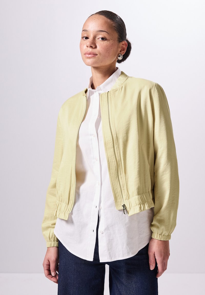 Giacca bomber gialla chiara con polsini e orlo elasticizzati, realizzata in tessuto liscio. Indossata sopra una camicia bianca a maniche lunghe e jeans blu.