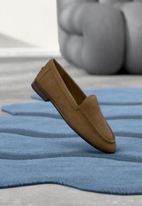 Mocassin en daim marron posé sur un tapis texturé bleu avec un canapé gris capitonné en arrière-plan.