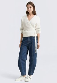 Vit stickad omlottkofta med långa ärmar och knytbälte, kombinerad med lösa blå denimjeans och vita sneakers.