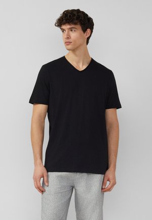 Mężczyzna w czarnej koszulce z krótkim rękawem i dekoltem w szpic oraz jasnoszarych spodniach stojący na jednolitym szarym tle.