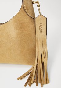 Sacs en daim beige présentant une silhouette incurvée, des coutures contrastées et une étiquette en cuir avec des accents en franges. Détails en métal doré inclus.