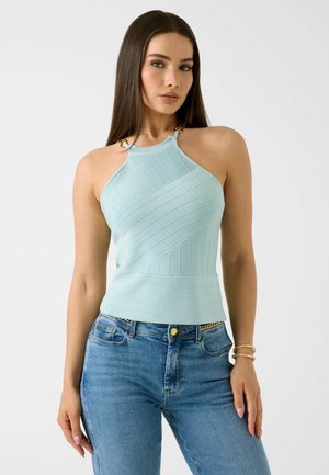 Femme aux longs cheveux foncés portant un haut sans manches côtelé bleu clair et un jean bleu, debout devant un fond blanc uni.