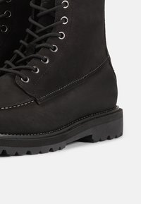 Bota de senderismo de cuero negro con punta redonda, ojales de metal y cordones negros. Presenta una suela texturizada con surcos prominentes y acentos de costura blanca.