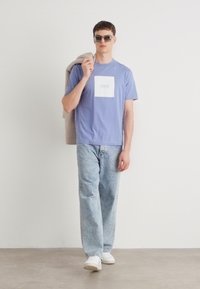 T-shirt lilás claro com um logótipo quadrado branco, jeans de ganga azul e ténis brancos. O modelo segura uma jaqueta bege sobre o ombro.