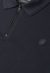 Polo en maille côtelée bleu marine foncé avec une fermeture éclair d’un quart et un écusson rond noir avec logo sur la poitrine.