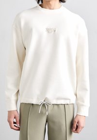 Sudadera de color crema claro con cuello redondo, mangas largas y dobladillo ajustable con cordón. Presenta un sutil logo bordado en el pecho.