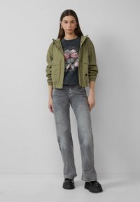 Olijfgroene hoodie jas, grijs T-shirt met een vlinderprint, lichtgrijze uitlopende spijkerbroek en zwarte platform schoenen.
