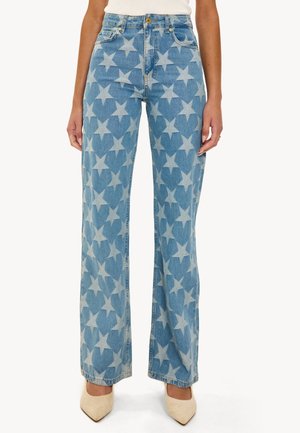 Jean en denim bleu à jambes larges avec motifs étoiles clairs sur toute la surface, associé à des talons beiges à bout pointu et un haut blanc.