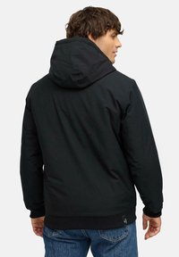 Svart huvanjacka med en slät textur, elastiska manschetter och en synlig logotagg längst ner. Har en avslappnad passform och minimalistisk design.
