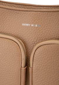Detailní záběr texturované hnědé kožené tašky se dvěma paralelními zipovými kapsami a nápisem "GERRY WEBER" ve stříbrném písmu.