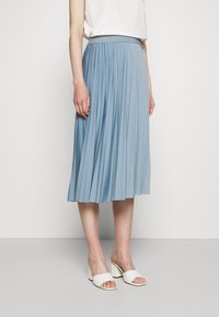 Hellblauer plissierter Rock mit elastischem Bund und strukturierter Oberfläche, kombiniert mit weißen Sandalen mit Absatz und einem schlichten weißen Oberteil.