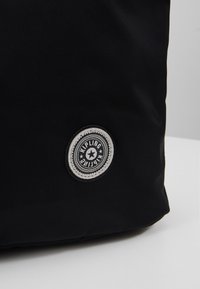 Sac en tissu noir avec un écusson rond noir et blanc du logo Kipling, étoile au centre, près du coin inférieur.