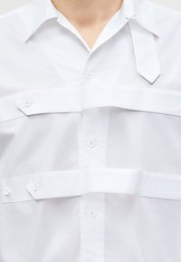 Λευκό πουκάμισο με γιακά, με πολλαπλές οριζόντιες υφασμάτινες λωρίδες που έχουν κουμπώματα στην περιοχή του στήθους.