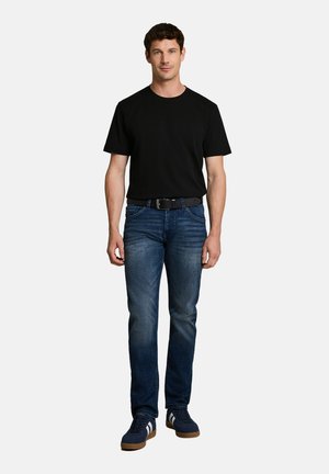 Man staat, draagt een zwart T-shirt, blauwe spijkerbroek met een zwarte riem en blauwe sneakers met witte strepen tegen een witte achtergrond.