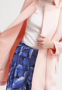 Chaqueta estructurada de color rosa claro con cierre de cremallera, llevada sobre una blusa blanca y una falda azul con estampado floral, que muestra una tela lisa y líneas limpias.