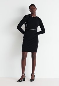 Μαύρο φόρεμα με μακριά μανίκια και λευκή περίγραμμα, εφαρμοστή σιλουέτα, μήκους μέχρι το γόνατο, με μαλακό ύφασμα, συνδυασμένο με μαύρες ψηλοτάκουνες γόβες και βραχιόλια αστραγάλου.