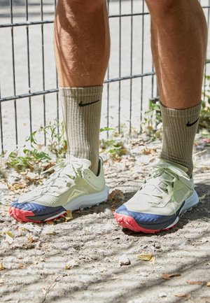 Beine mit beigen Nike-Socken und weißen Sportschuhen mit roten und blauen Akzenten, die auf sandigem Boden in der Nähe eines Metallzauns stehen.