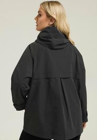 Veste de pluie noire avec capuche, présentant une coupe décontractée, un dos plissé et des poignets sécurisés. Fabriquée à partir d'un matériau léger et résistant à l'eau.