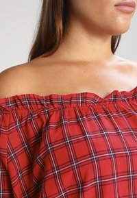 Femme portant un haut rouge à épaules dénudées avec un motif à carreaux noir et blanc, dévoilant le cou, les épaules et une partie du visage.