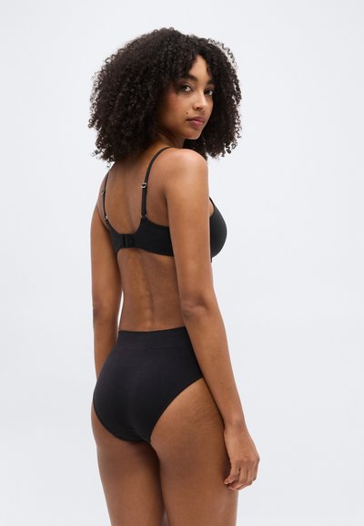 Γυναίκα με σγουρά μαλλιά που φοράει μαύρο σουτιέν και μαύρα εσώρουχα με ψηλή μέση, γυρισμένη ελαφρώς για να δείξει την πλάτη της και κοιτάζοντας πάνω από τον ώμο της.