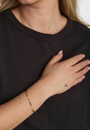 Czarna bawełniana koszulka z okrągłym dekoltem, noszona na ręce z złotą bransoletką i tatuażem na nadgarstku przedstawiającym stylizowaną literę.