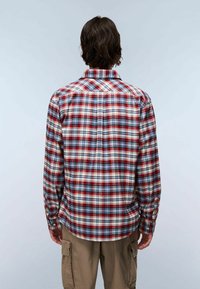 Uomo che indossa una camicia a maniche lunghe a quadri rossi, bianchi e blu e pantaloni cargo beige, in piedi di spalle contro uno sfondo semplice.