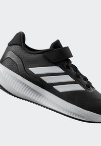 Chaussure de sport noire avec un dessus texturé, dotée de trois rayures blanches, d'une sangle de sécurité, et d'une semelle intermédiaire blanche flexible avec des motifs de traction.