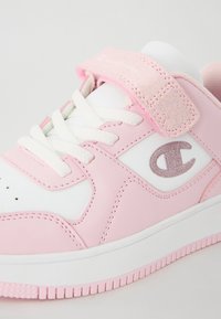 Baskets sportives roses et blanches avec une sangle en Velcro rose texturée, des lacets blancs et un accent de logo pailleté. Matériau synthétique lisse.