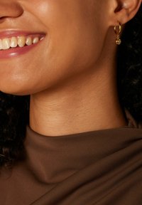 Goldene Creole mit einem kleinen hängenden Edelstein, vor glattem, braunem Stoff und strukturierter Haut.