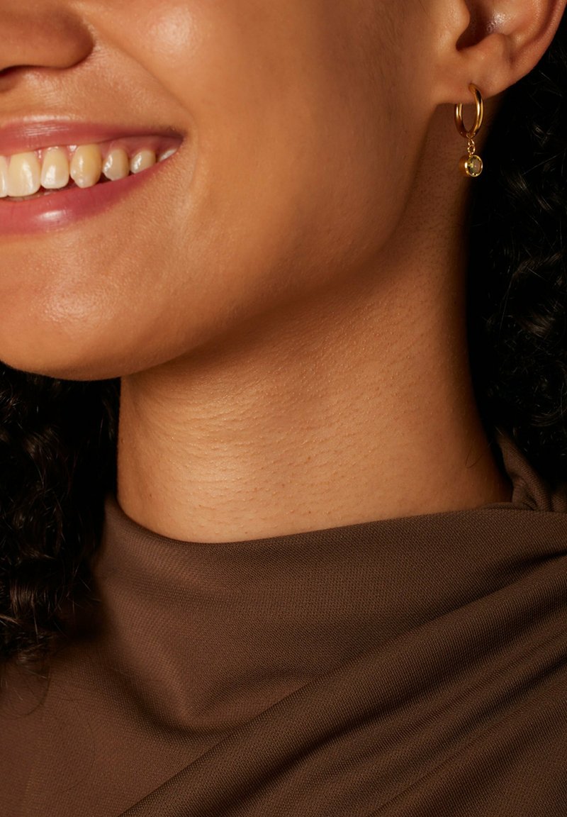 Goldene Creole mit einem kleinen hängenden Edelstein, vor glattem, braunem Stoff und strukturierter Haut.