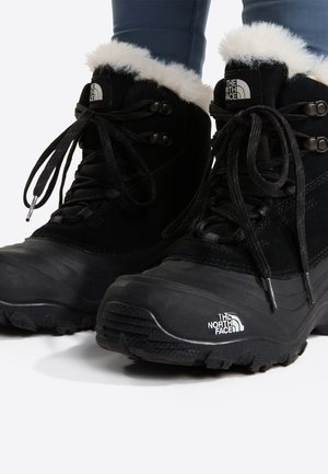 Черни зимни ботуши The North Face с кожа и връзки, носени с тъмни панталони на бял фон.