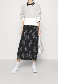 Maglione grigio e bianco a maglia sopra un abito maxi nero con figure in linea bianca, abbinato a sneakers bianche e una borsa testurizzata.