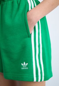Grønne bomullsshorts med hvite sidestriper og et brodert Adidas-logo nederst til venstre. Har sidelommer.