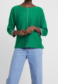 Groene gebreide trui met een ronde hals, grijze manchet en voornaaddetail. Gedragen met lichtgewassen jeans, met een relaxte pasvorm.