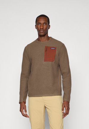 Brown textured sweater with a chest pocket in rust. Features a round neckline and long sleeves, paired with light beige pants.